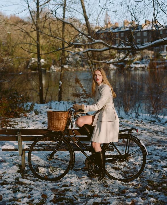 A woman sits on a bicycle with a basket in a snowy park next to a body of water and trees.