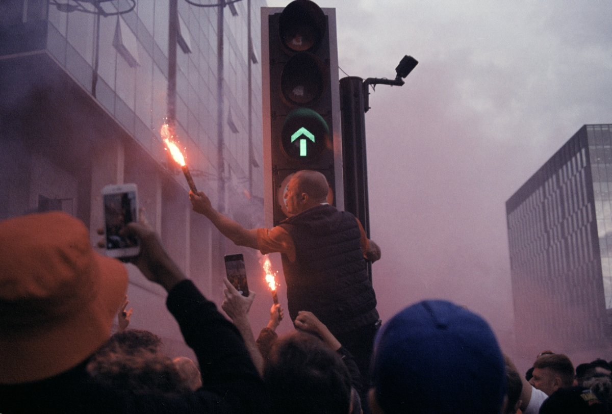 A man holds a flare next to a traffic light with an arrow while people film with their phones.