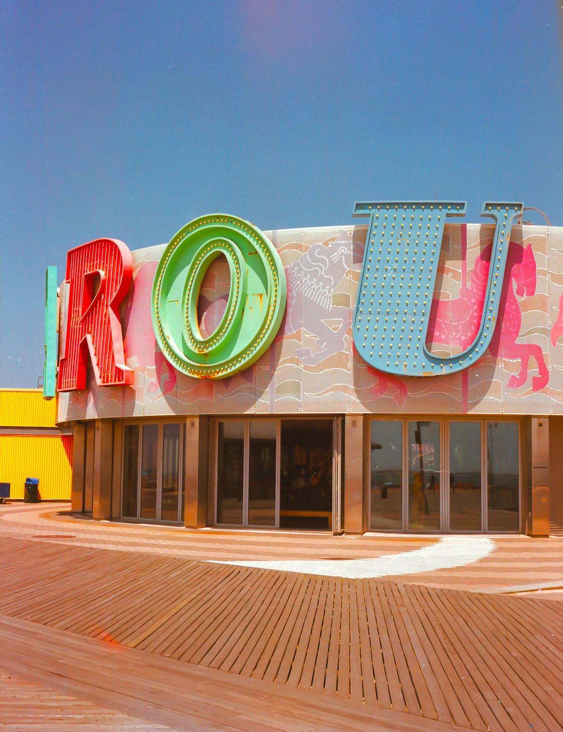 A building with large colorful letters spelling "ROU" stands on a wooden boardwalk under a clear blue sky.