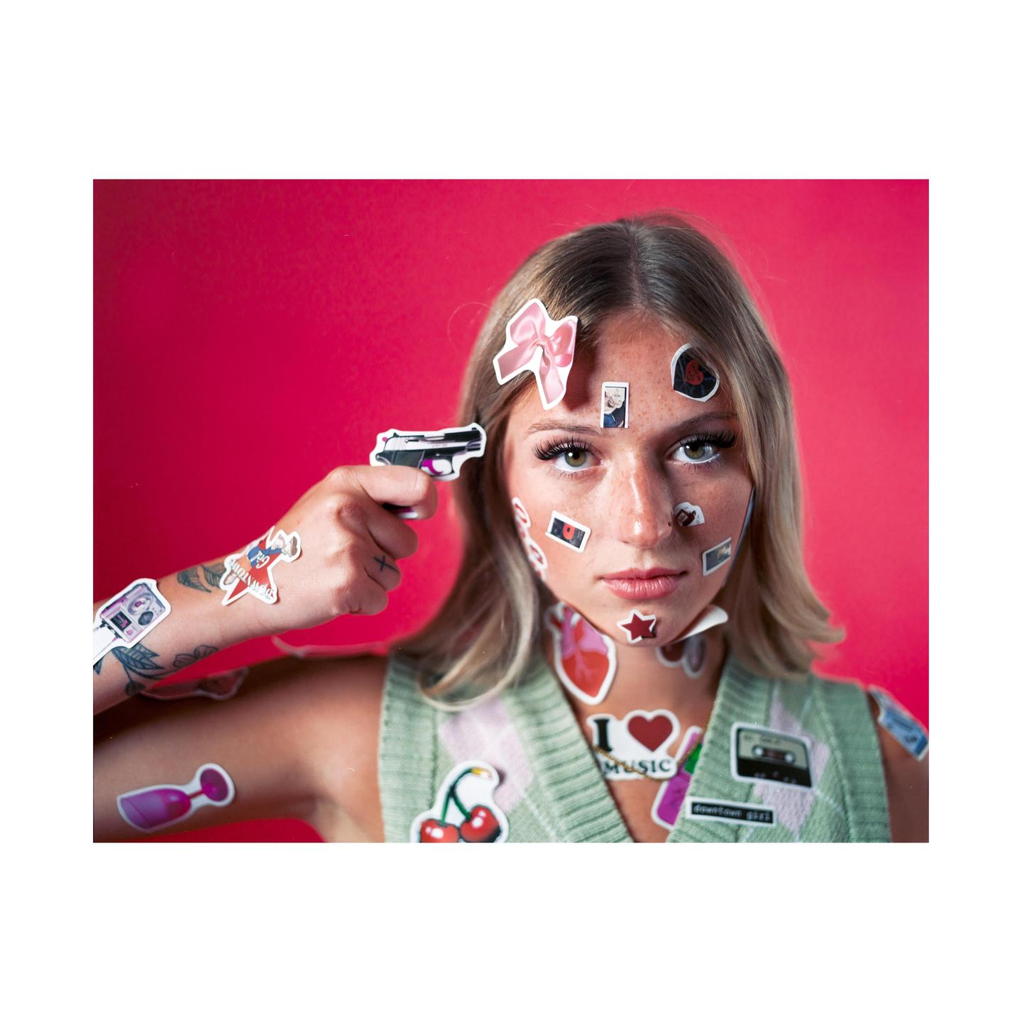 A woman with stickers on her face and body holds a toy gun to her head.