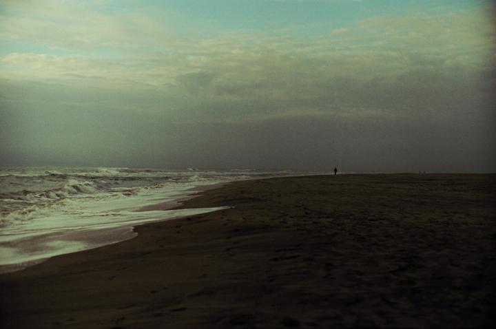 A lone figure walks along a dark beach with waves crashing on the shore under a cloudy sky.