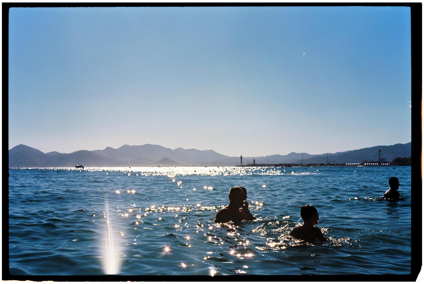 Three people are swimming in the sea with mountains and a pier in the background.