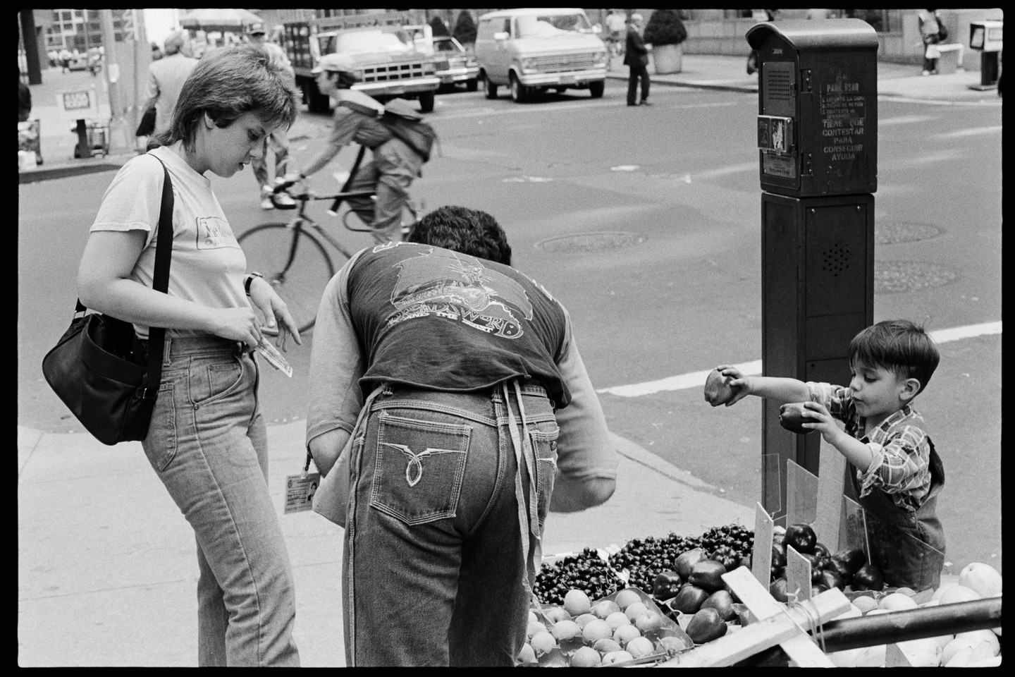 A woman, man, and child are at a street fruit stand in a city.