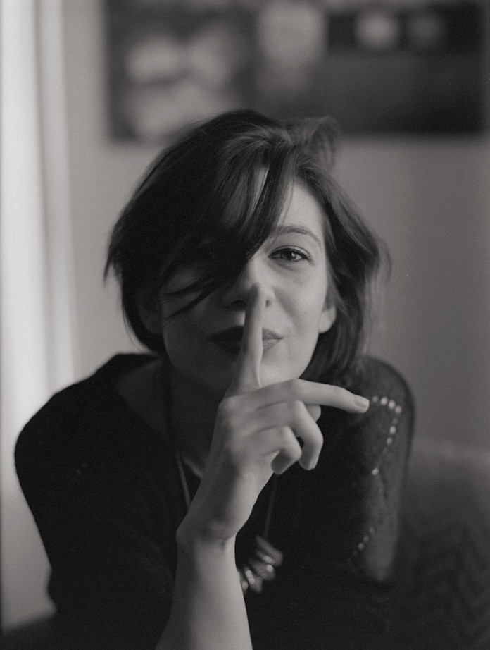 A woman with dark hair covering one eye holds a finger to her lips.