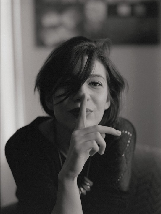 A woman with dark hair covering one eye holds a finger to her lips.