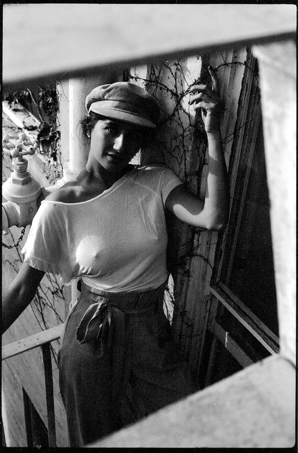 A woman in a cap and t-shirt stands on a balcony with a vine-covered wall behind her.