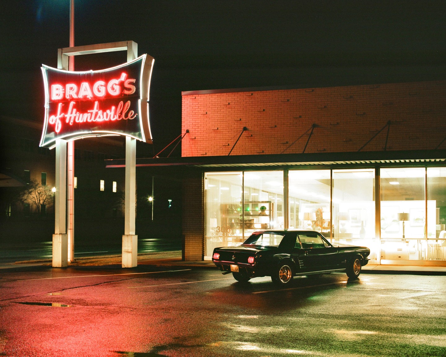 A black car is parked in front of a building with a neon sign that reads "Bragg's of Huntsville" at night.