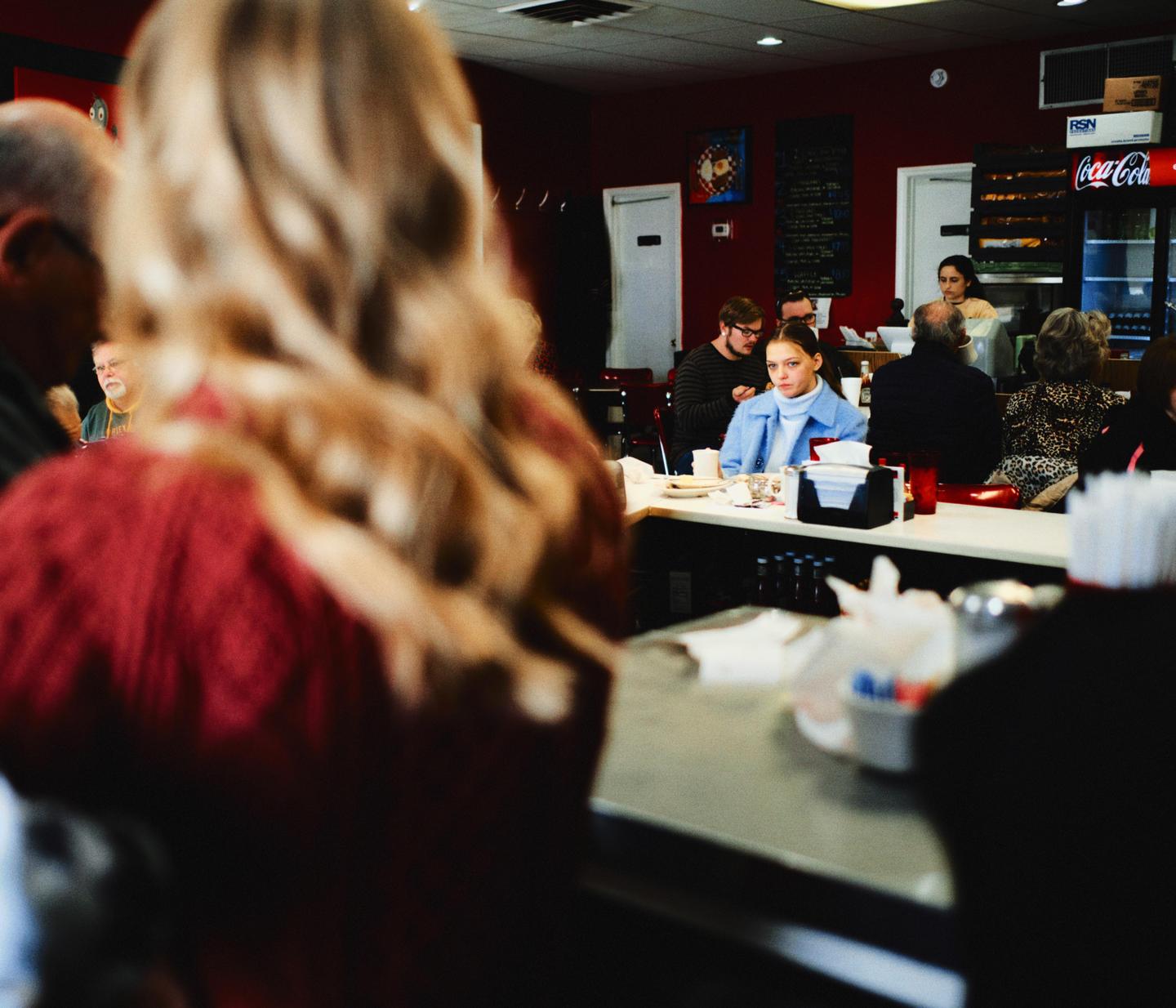 A woman in a blue jacket sits at a diner counter, looking towards the viewer.