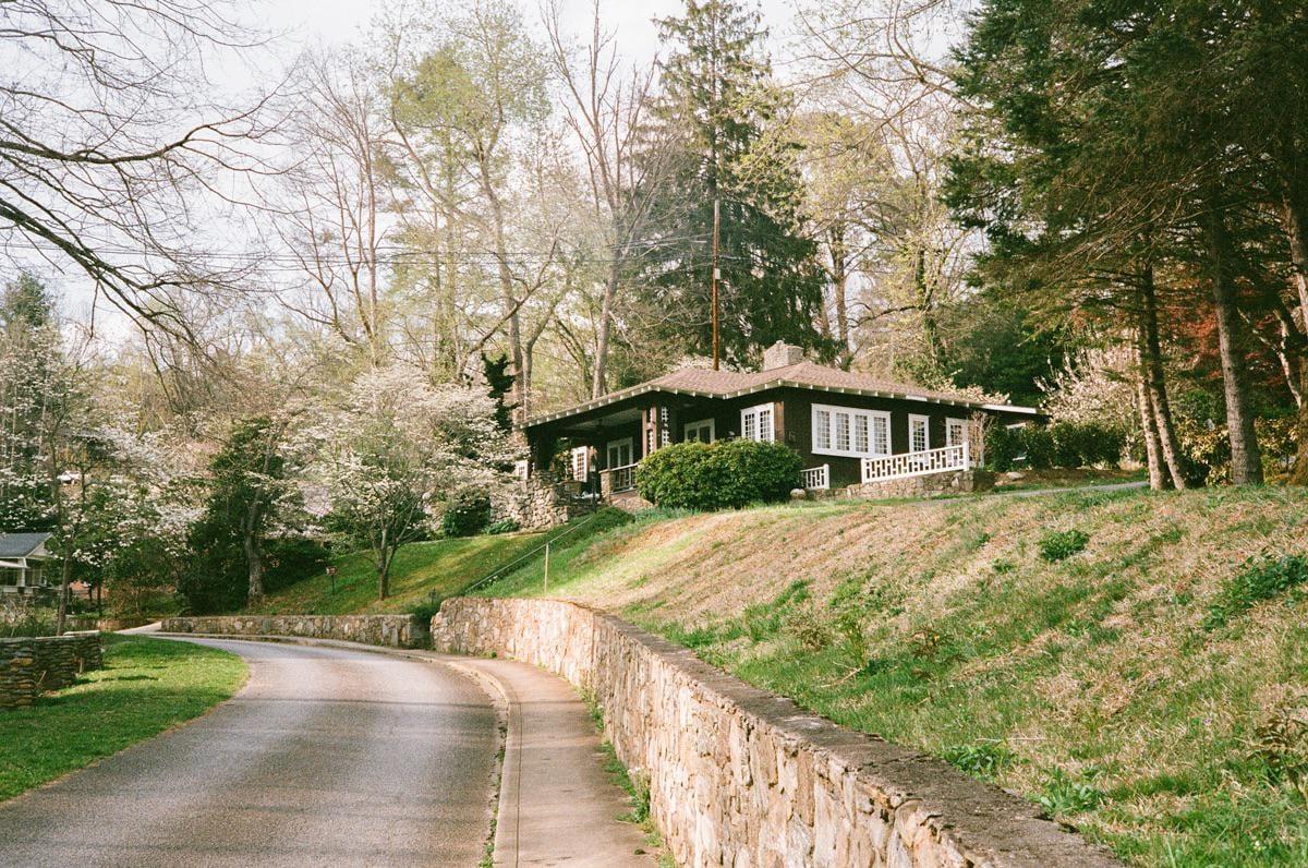 A winding road and stone wall lead up a grassy hill to a house surrounded by trees.