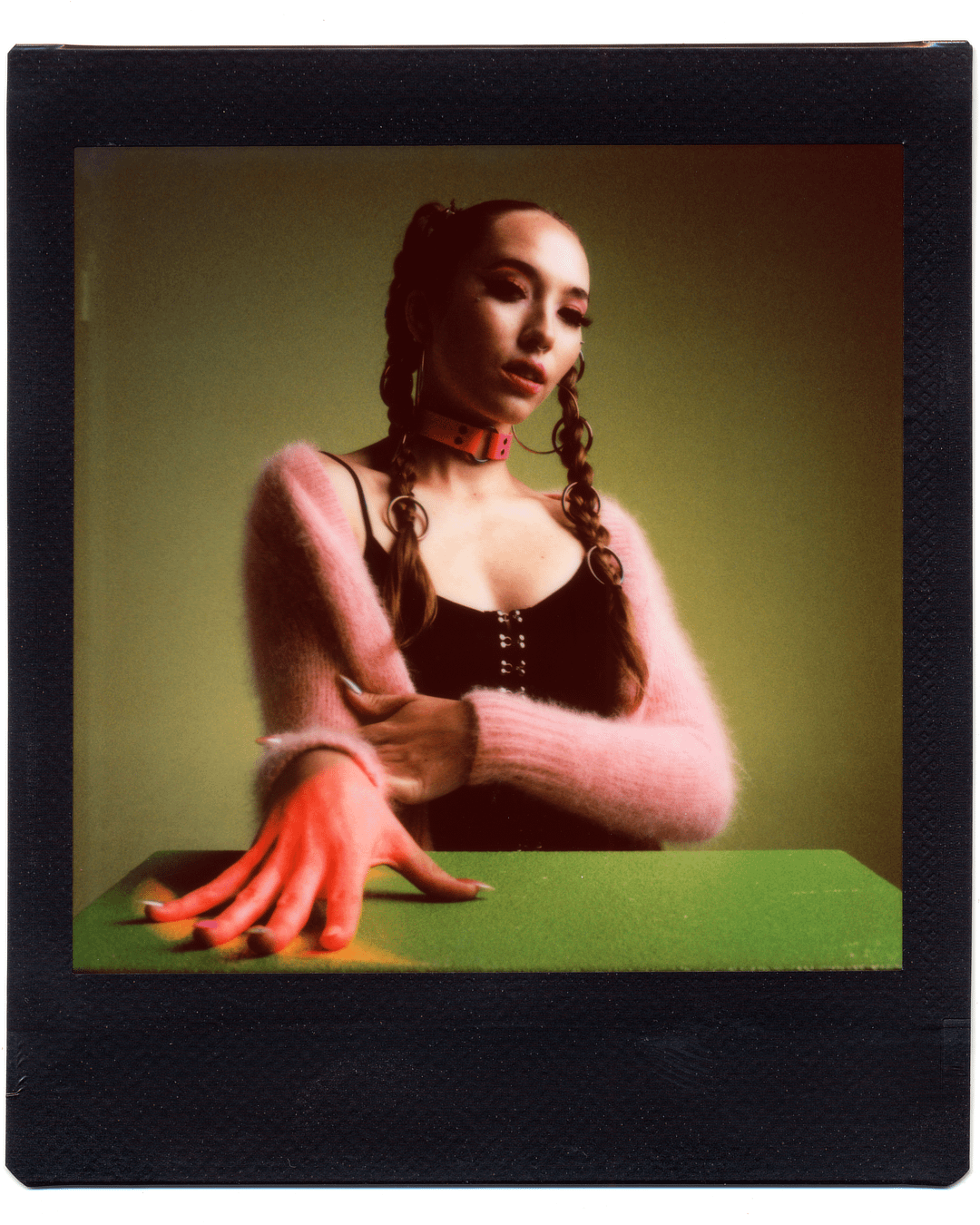 A woman with braided hair and a pink fluffy sweater poses with one hand on a green surface.