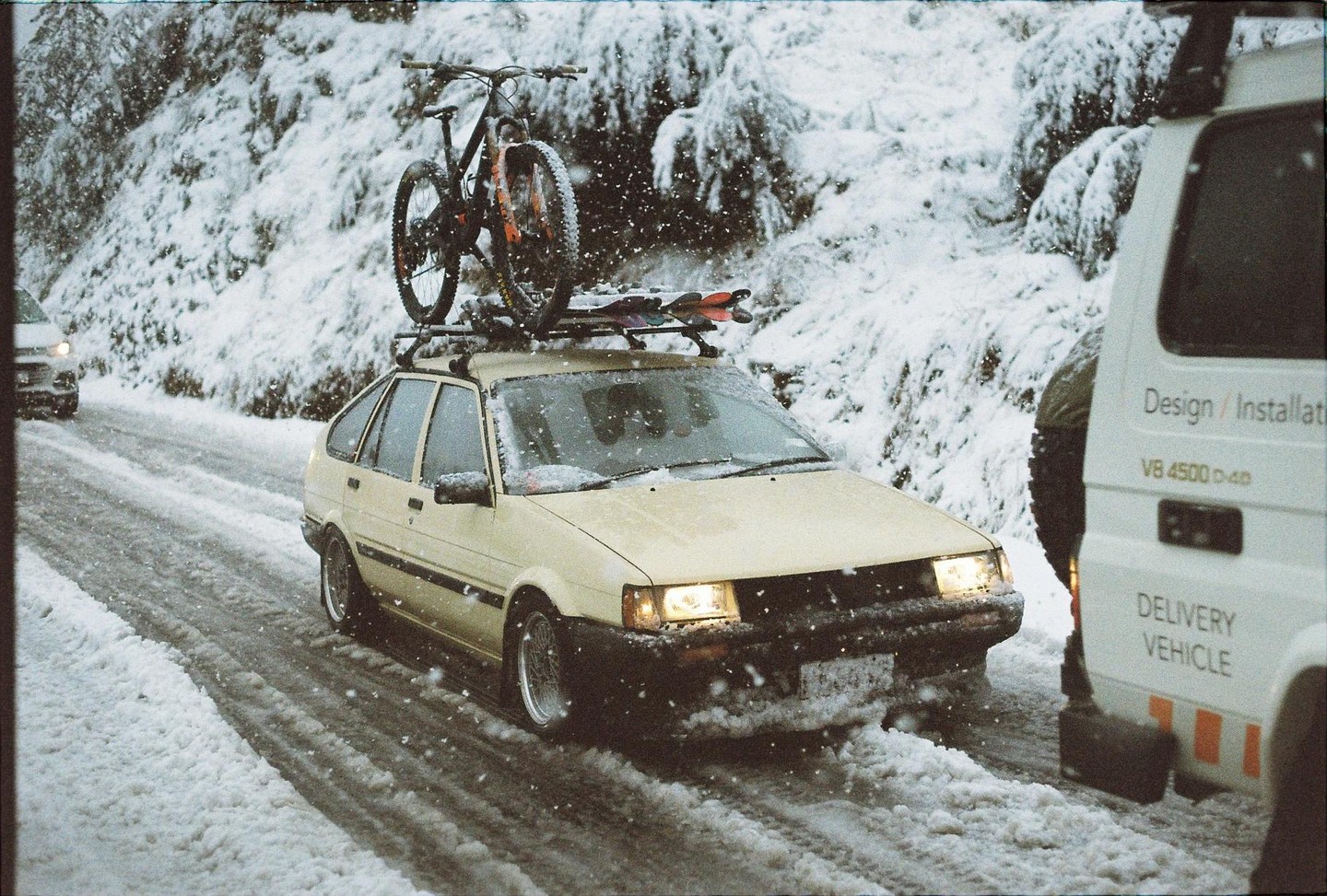A beige car with a bicycle and skis on its roof rack drives on a snowy road next to a white delivery vehicle.