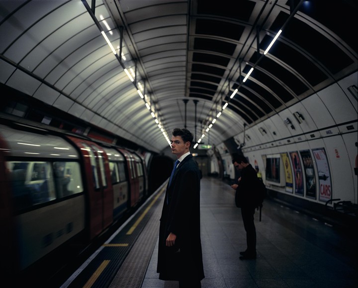A man in a coat and tie stands on a subway platform as a train passes by.