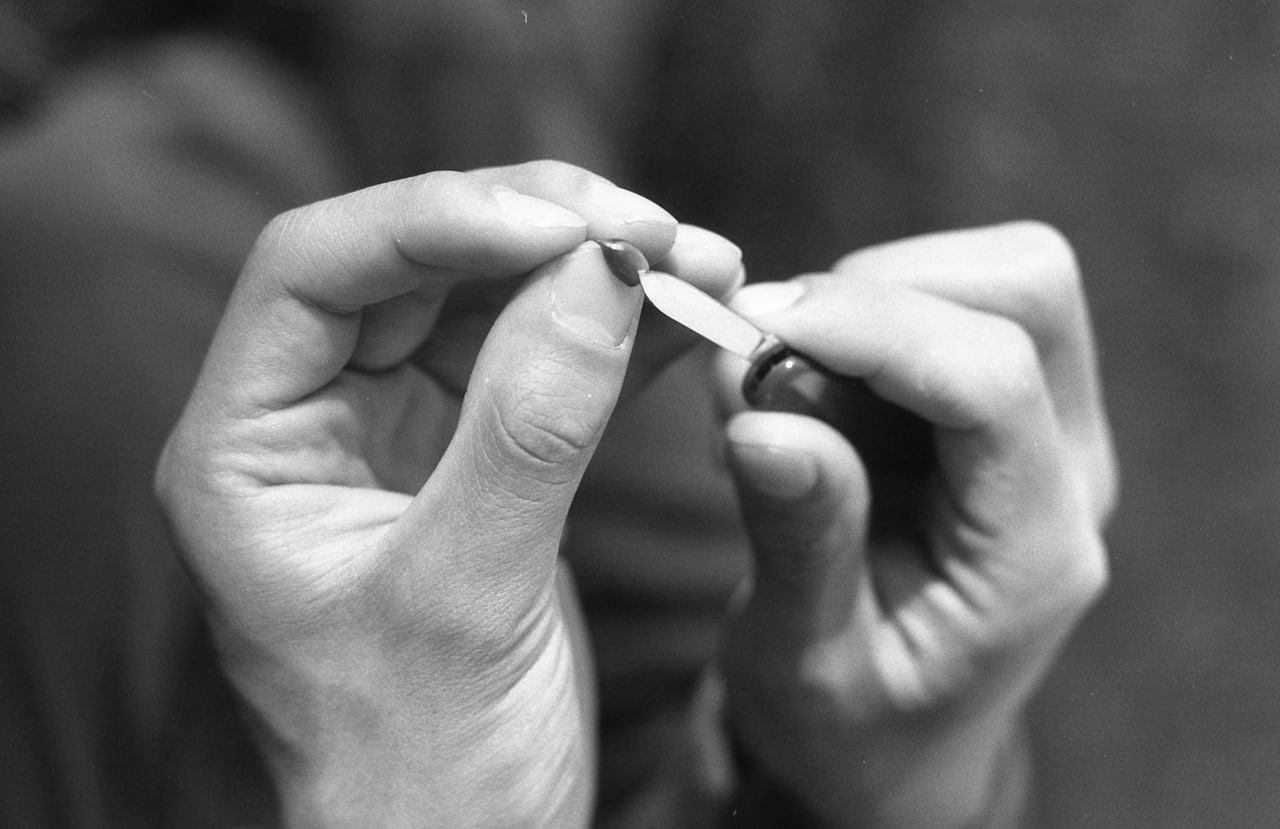 Two hands are shown in close-up, one holding a small object and the other using a tiny knife to scrape it.