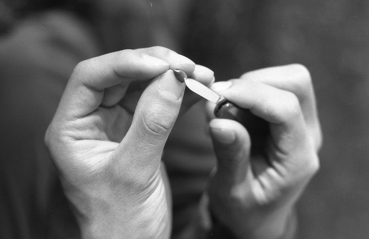 Two hands are shown in close-up, one holding a small object and the other using a tiny knife to scrape it.