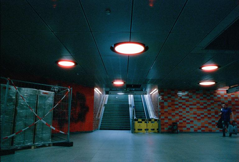 A man walks through an underground station with red lights on the ceiling and a tiled wall.