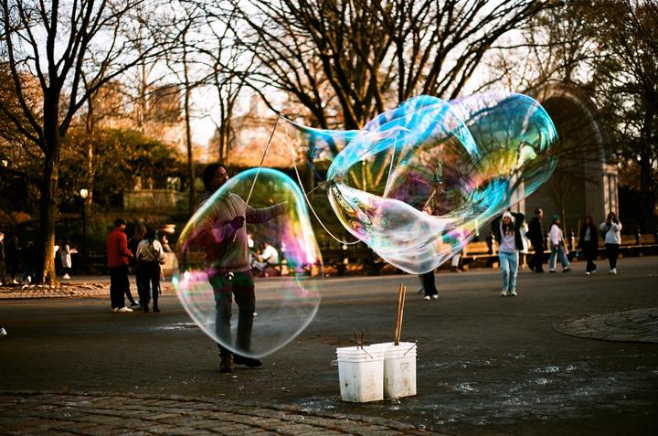 A man creates large, colorful bubbles in a park with bare trees and other people in the background.