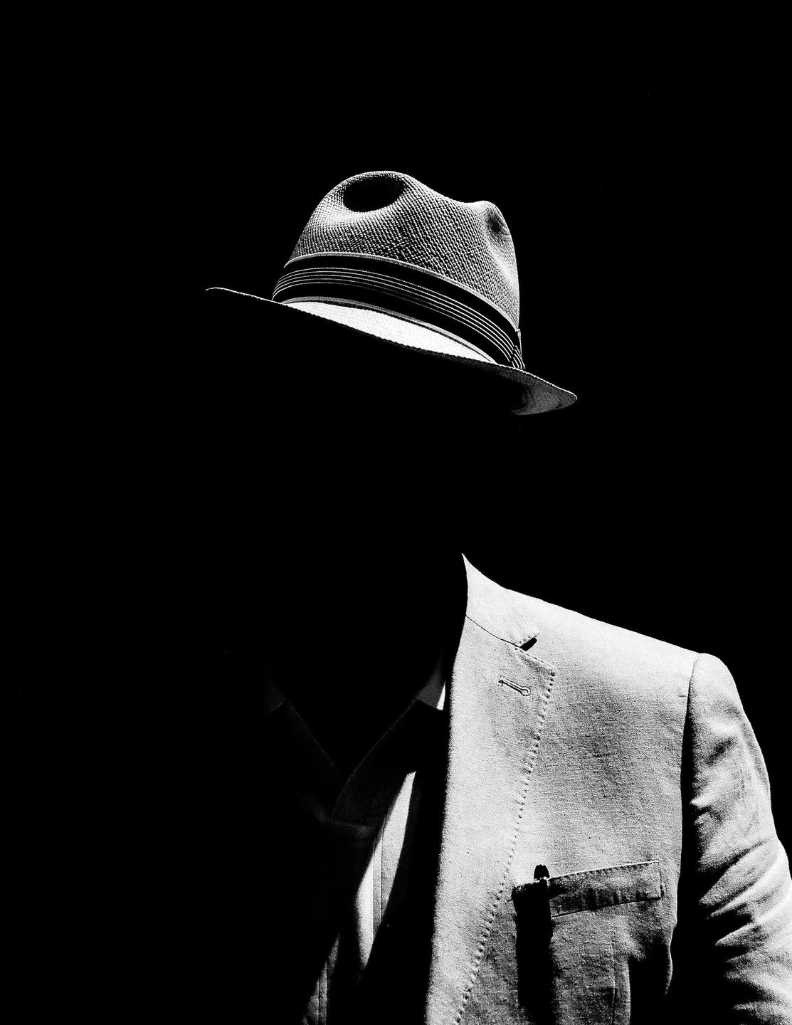 A man in a fedora and suit jacket is visible in strong contrast against a dark background.