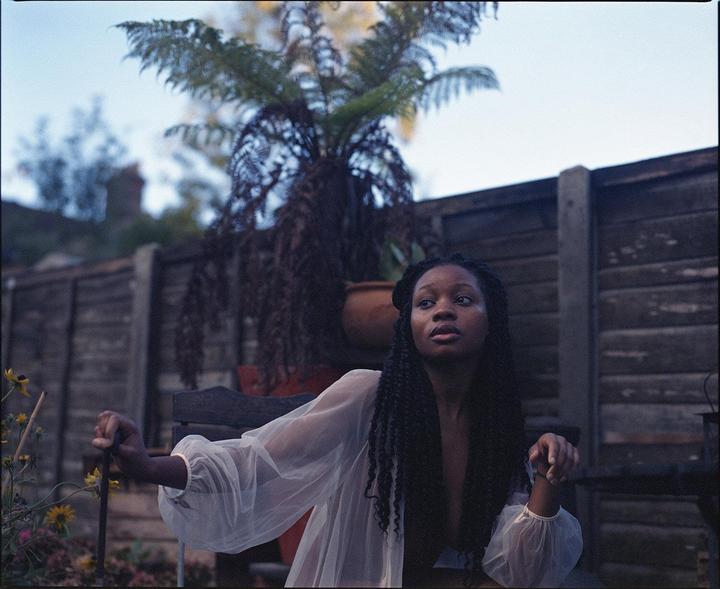 A woman with long braids sits outdoors, looking up and to the right, with a wooden fence and potted plant in the background.