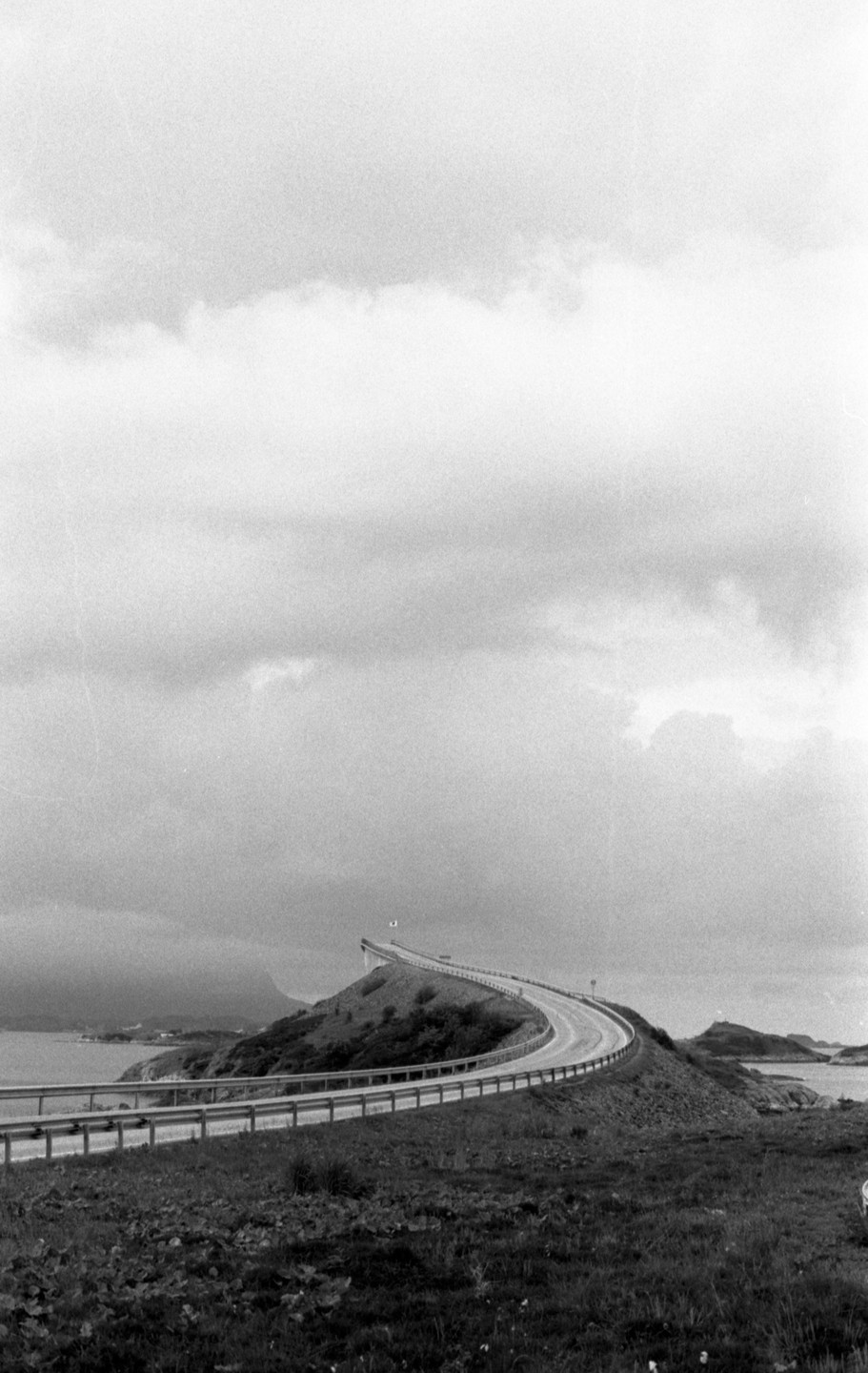 A winding road curves over a small hill next to a body of water under a cloudy sky.