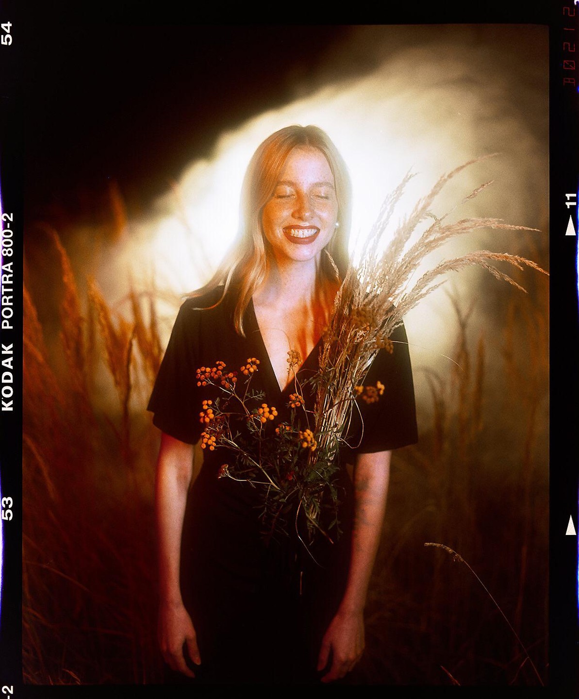 A woman with closed eyes and a wide smile holds a bouquet of dried grass and small orange flowers.