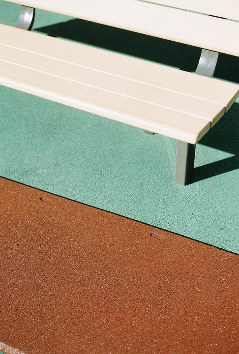 A light-colored bench casts a shadow on a green and brown textured ground.