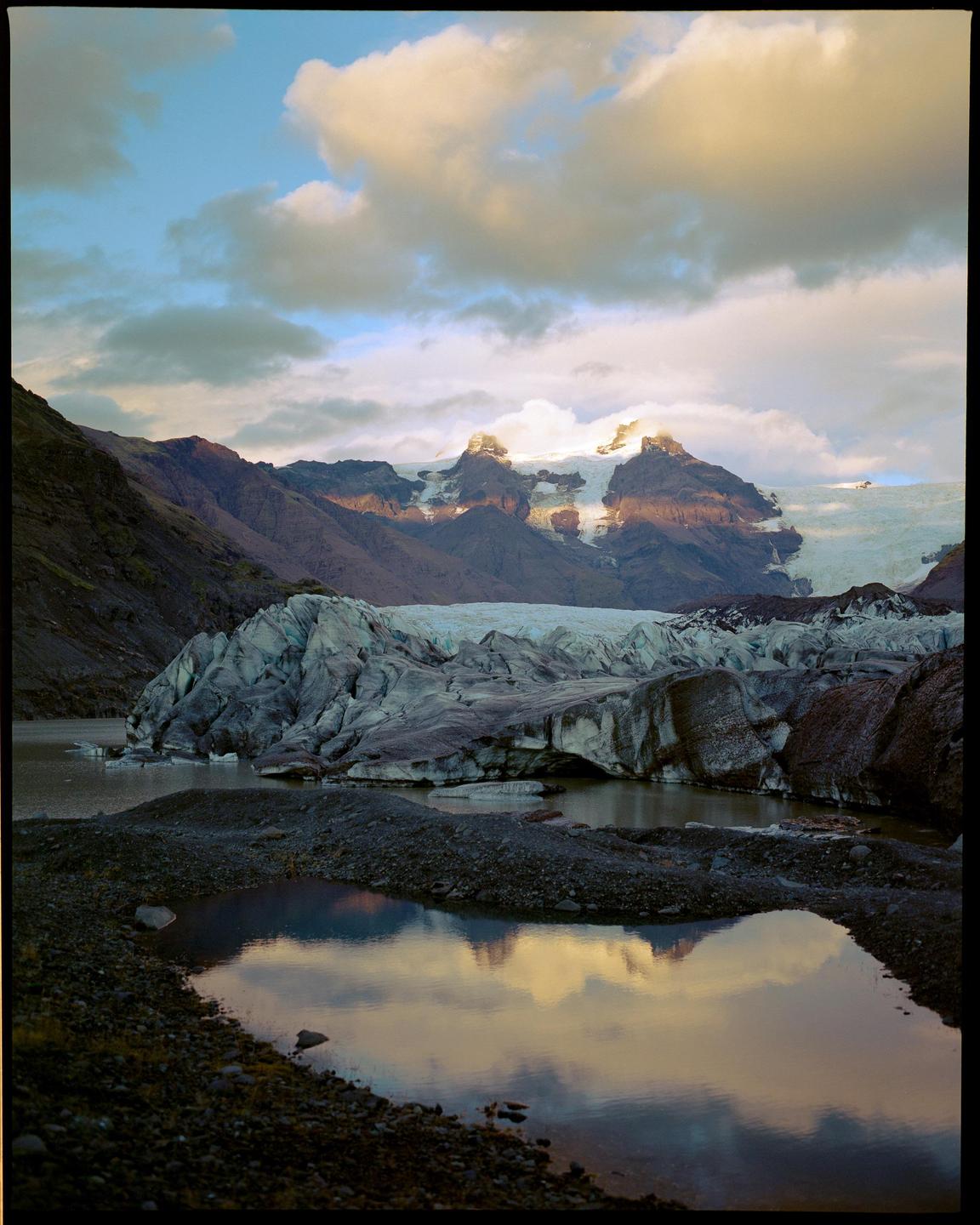 A glacier and mountains are reflected in a body of water under a cloudy sky.