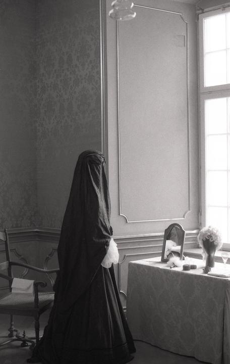 A person in a dark, flowing garment stands next to a dressing table with a mirror and a wig.