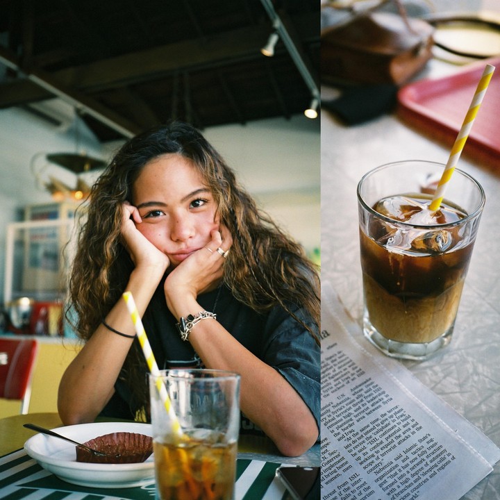 A woman with curly hair rests her chin on her hands at a table with a drink and a pastry, next to a glass of iced coffee on a newspaper.