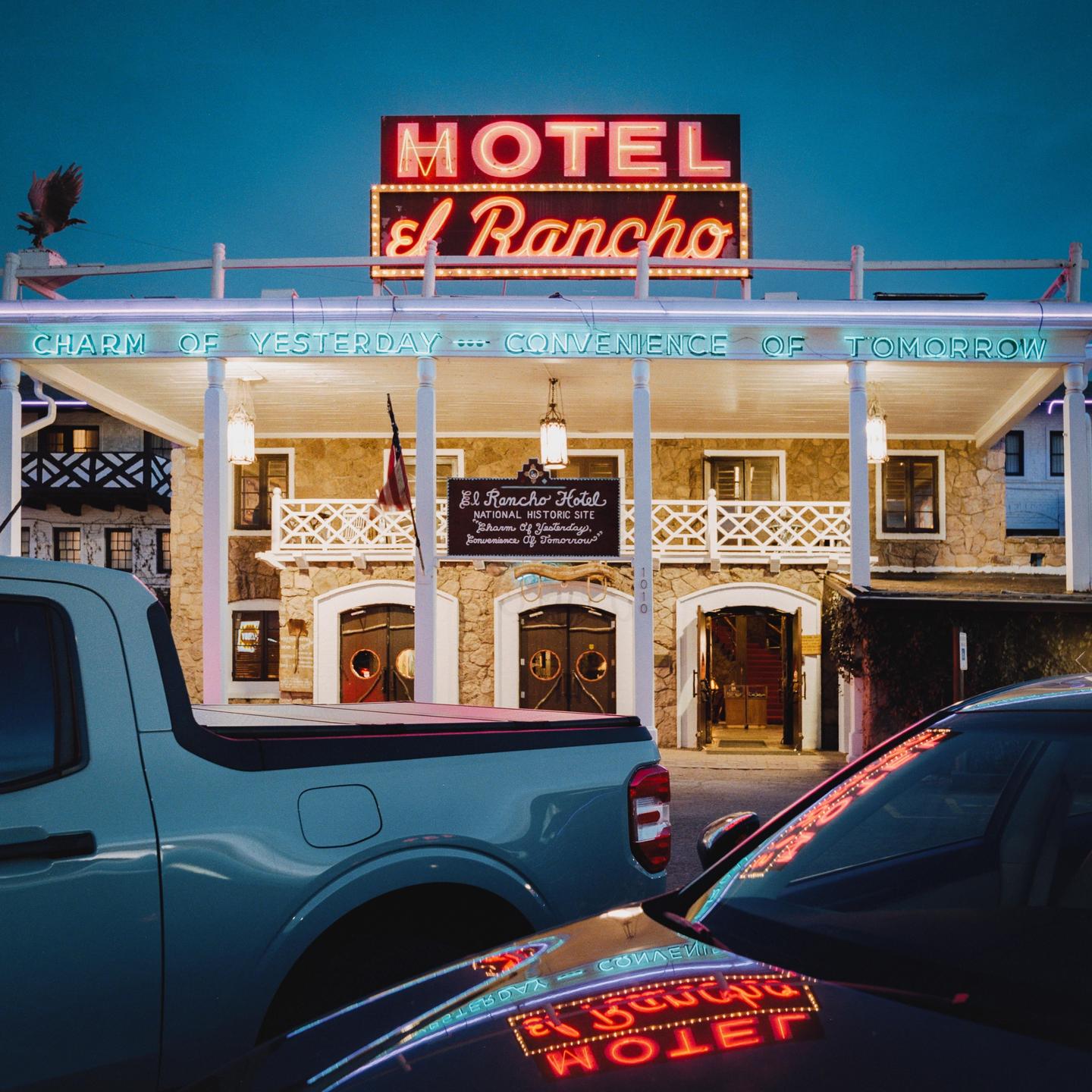 A neon sign for Hotel El Rancho is reflected in the hood of a car parked in front of the hotel at dusk.