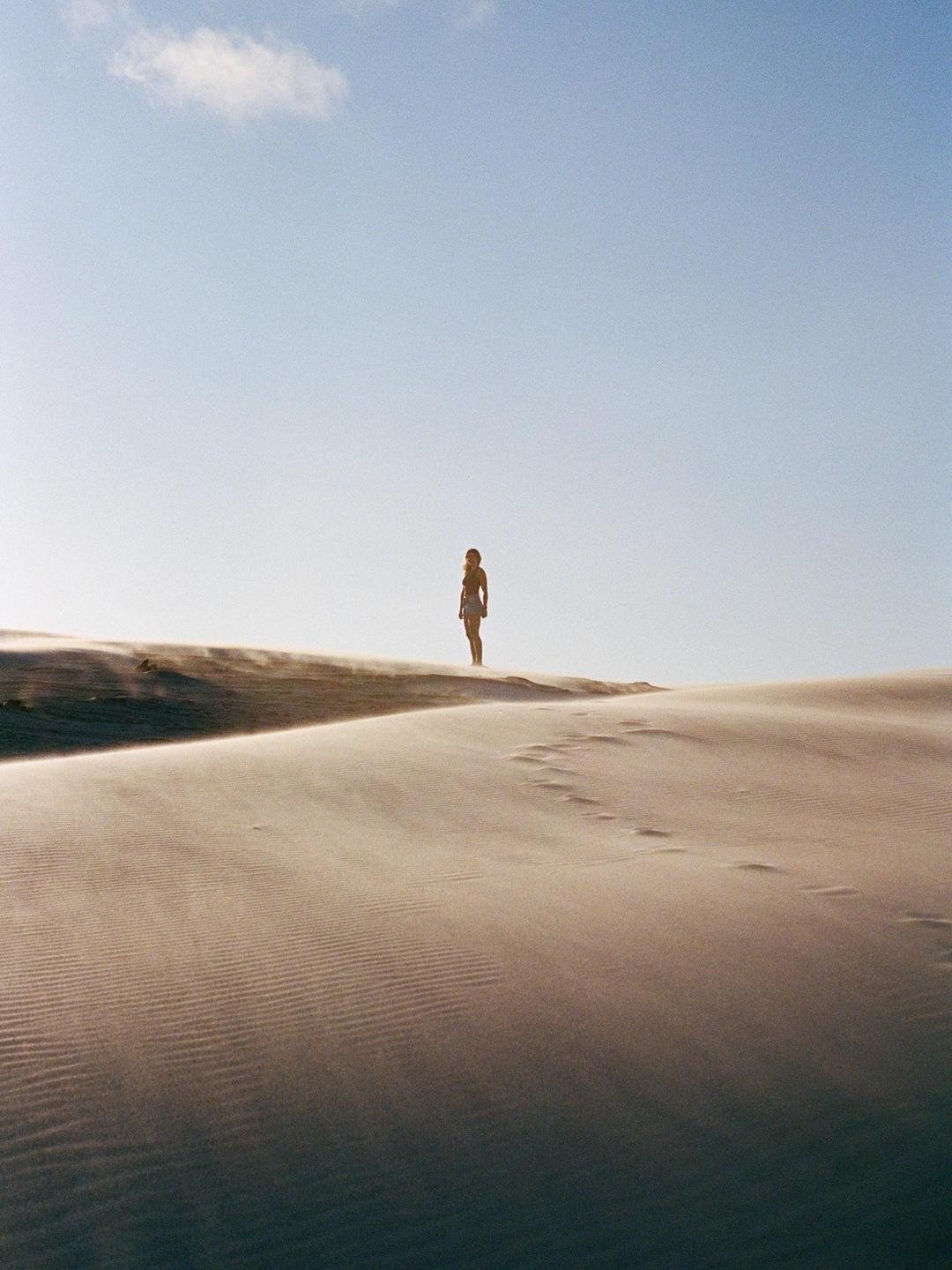A woman stands on a dune with sand blowing across the landscape under a clear sky.
