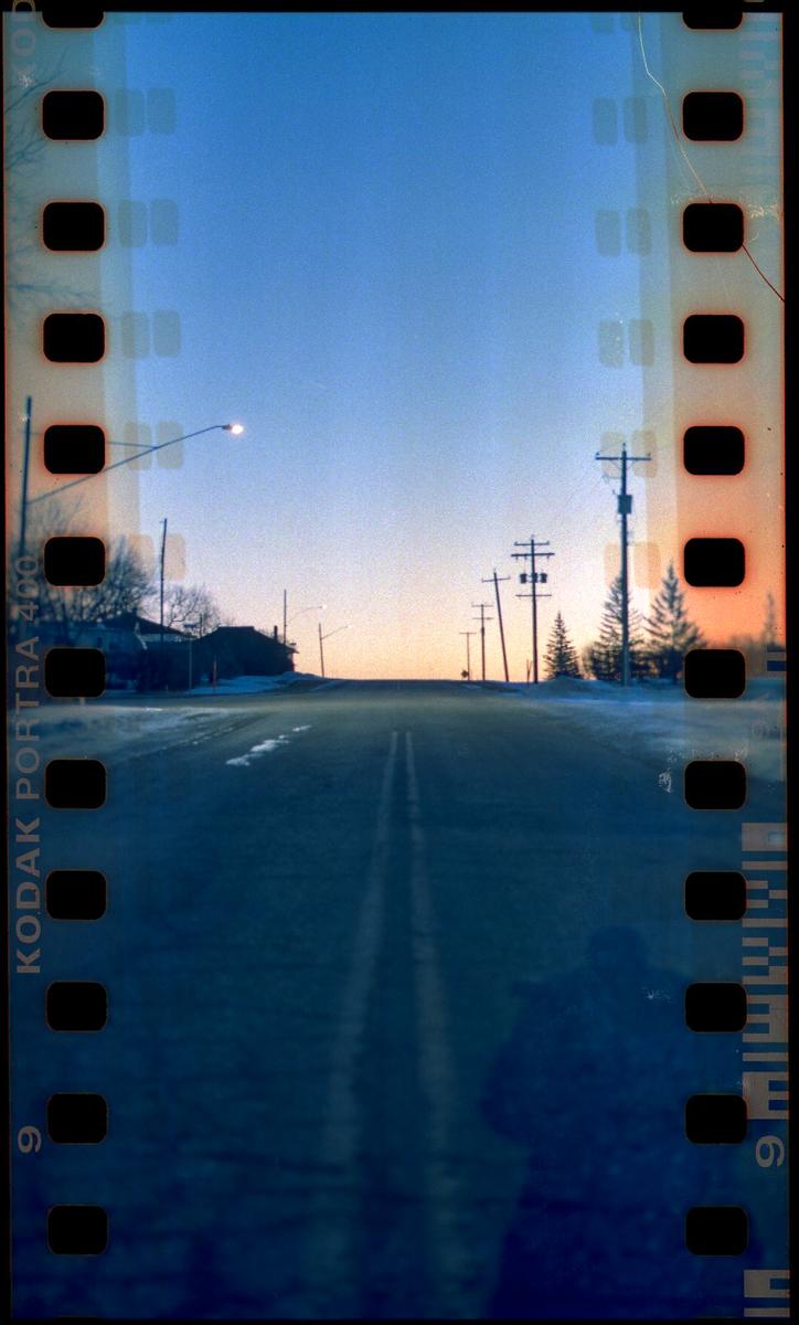 A road stretches into the distance under a sunrise sky, flanked by utility poles and snow-dusted trees.