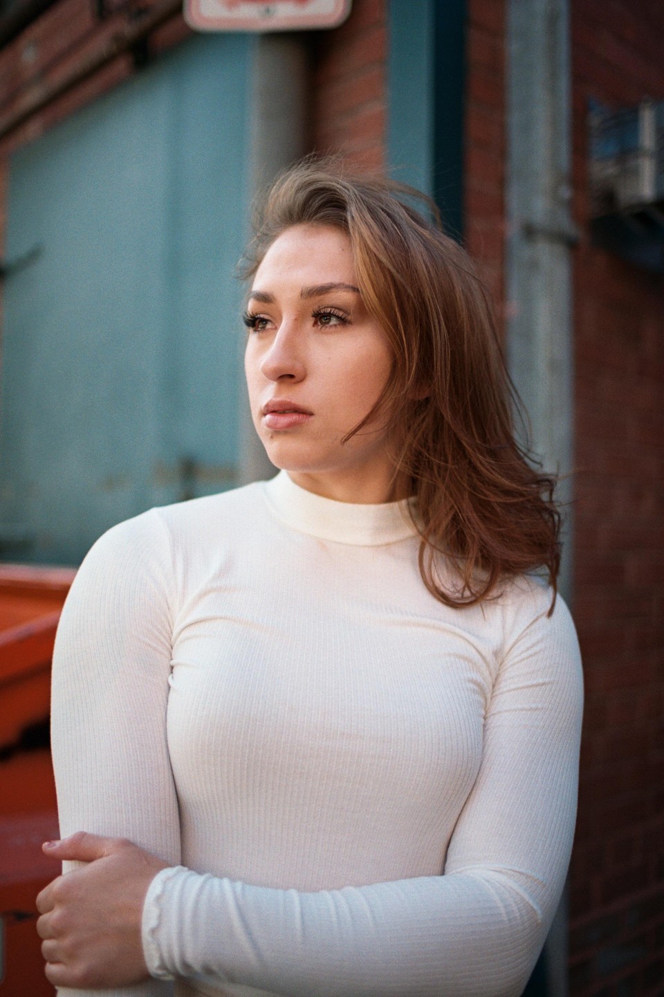 A woman with long hair and a white shirt stands outdoors with her arms crossed.