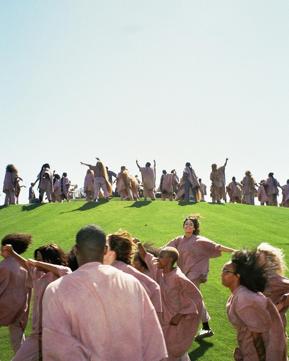 A large group of people in pink robes are gathered on a grassy hill under a clear sky.