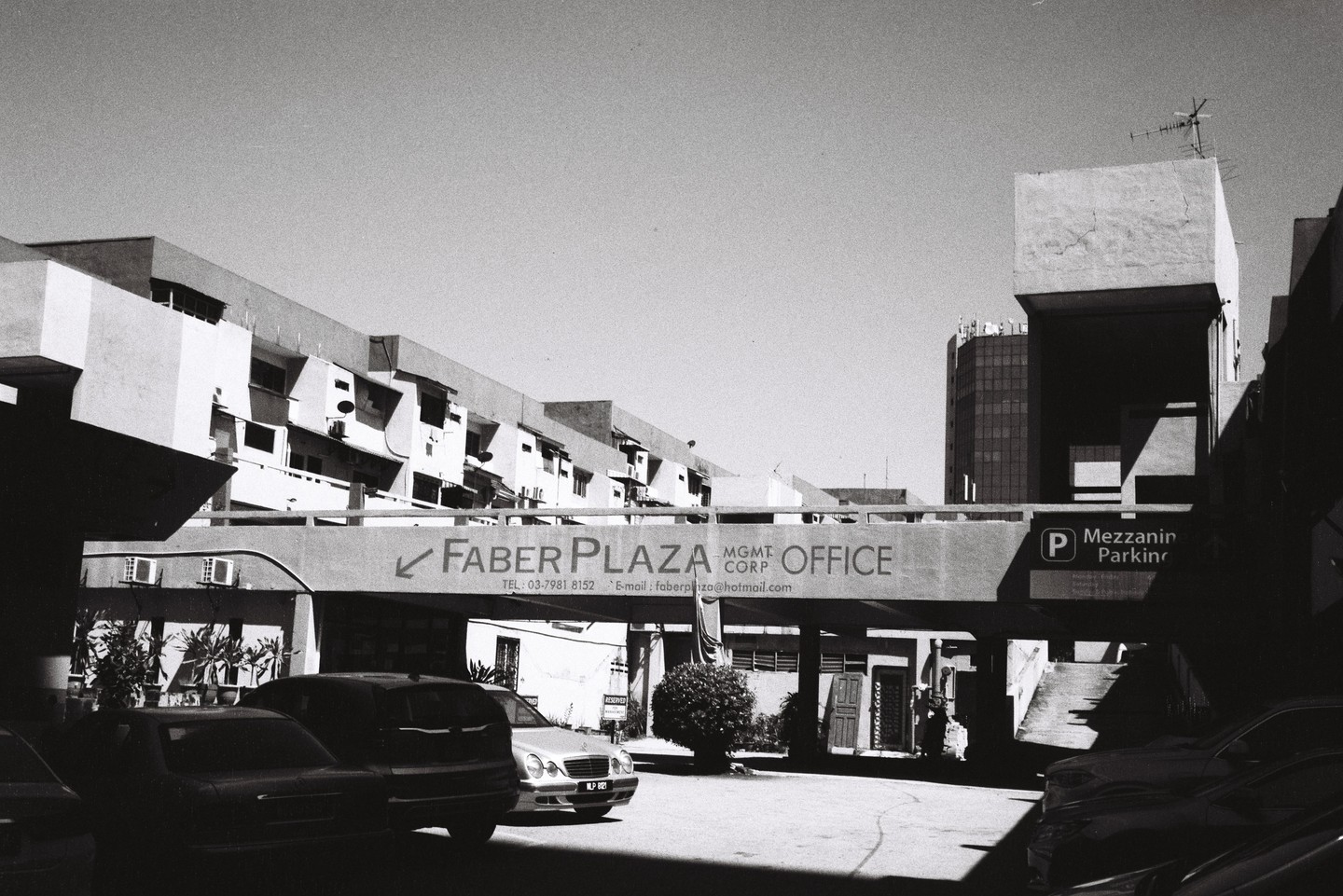 A black and white street photograph shows a parking lot with cars and buildings, including one with a sign for "Faber Plaza Office."