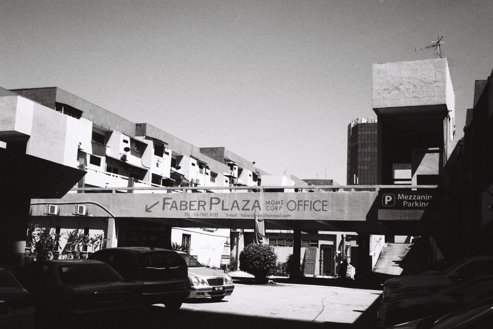A black and white street photograph shows a parking lot with cars and buildings, including one with a sign for "Faber Plaza Office."