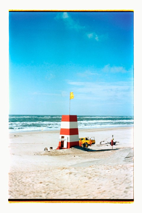 A red and white striped lifeguard tower stands on a sandy beach with a yellow flag flying from its top, next to a yellow pickup truck and the ocean.
