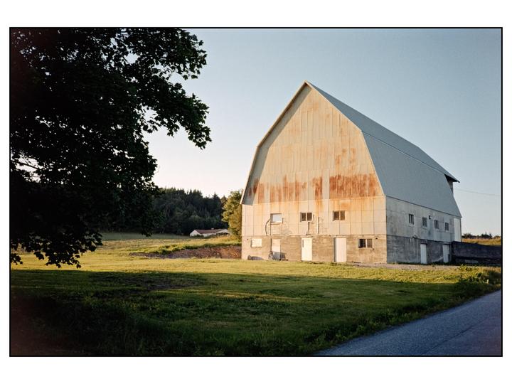 A large barn with a metal roof stands next to a field with a tree in the foreground.