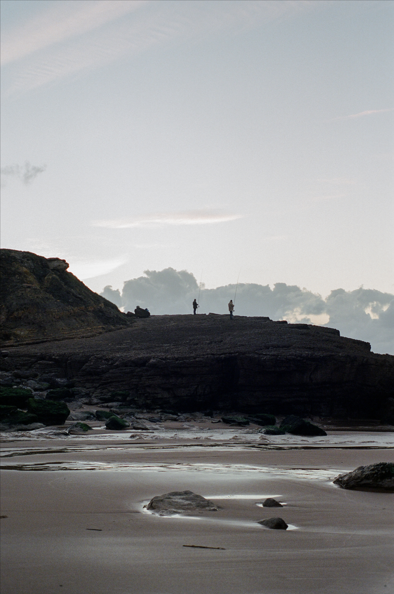 Two men fish from a rocky outcrop overlooking a sandy beach with tidal pools.