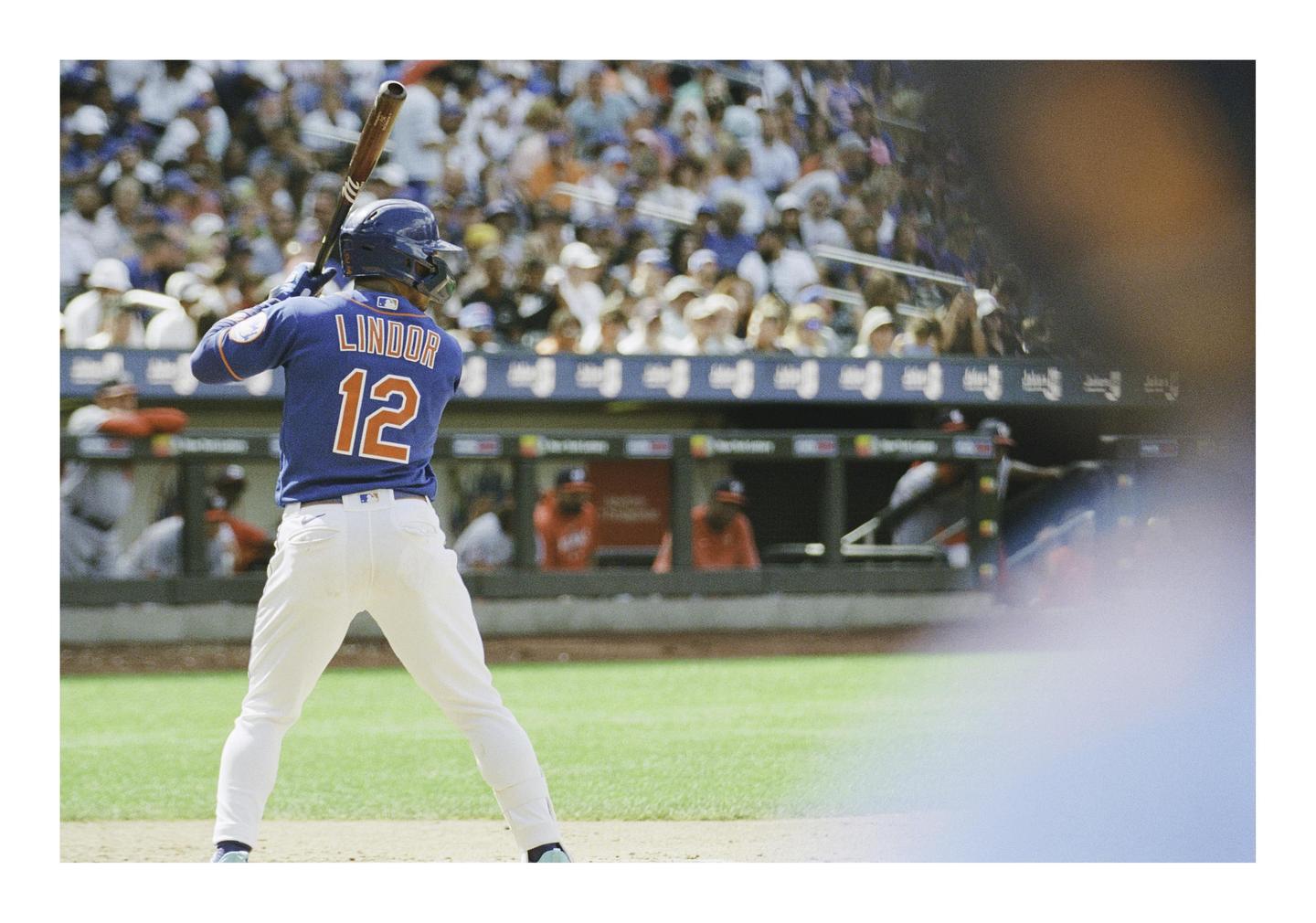 A baseball player in a blue jersey with the name "LINDOR" and number "12" on the back is at bat, facing away from the viewer, with spectators in the stands behind him.