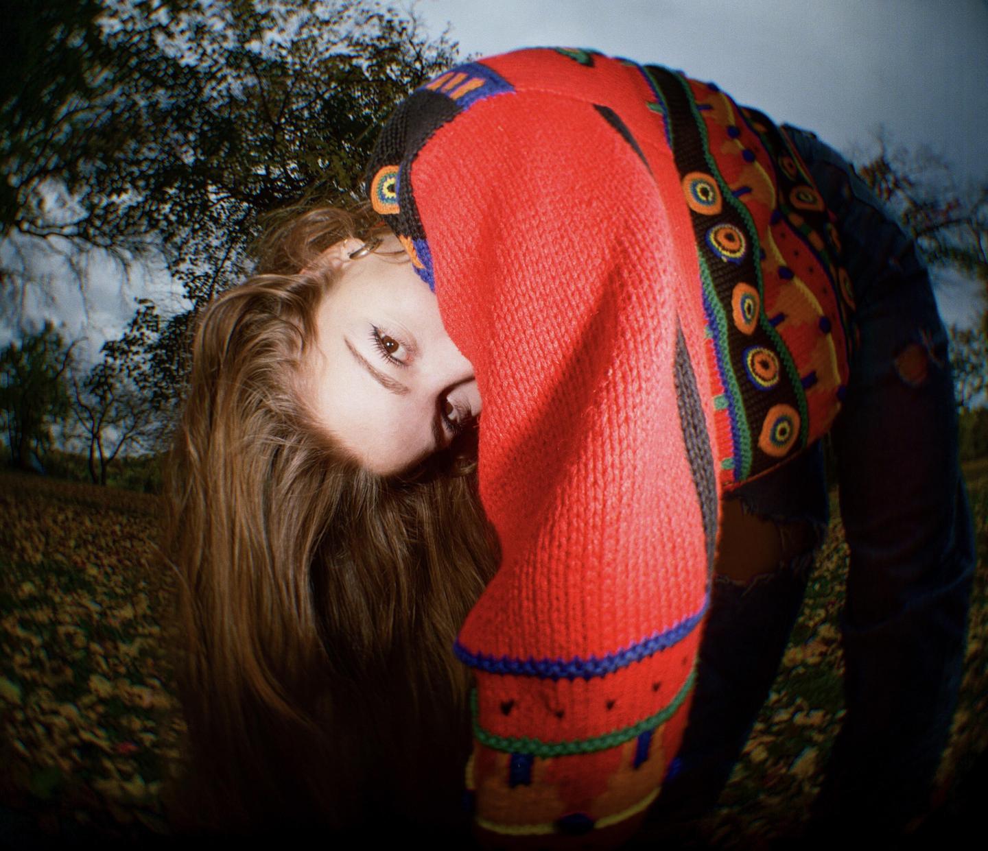 A woman in a colorful sweater is bending over with her face partially visible, surrounded by trees and fallen leaves.