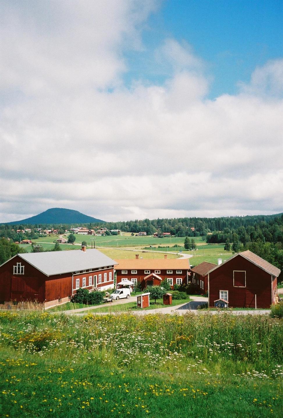 Red farm buildings are nestled in a green valley with a mountain in the distance under a cloudy sky.