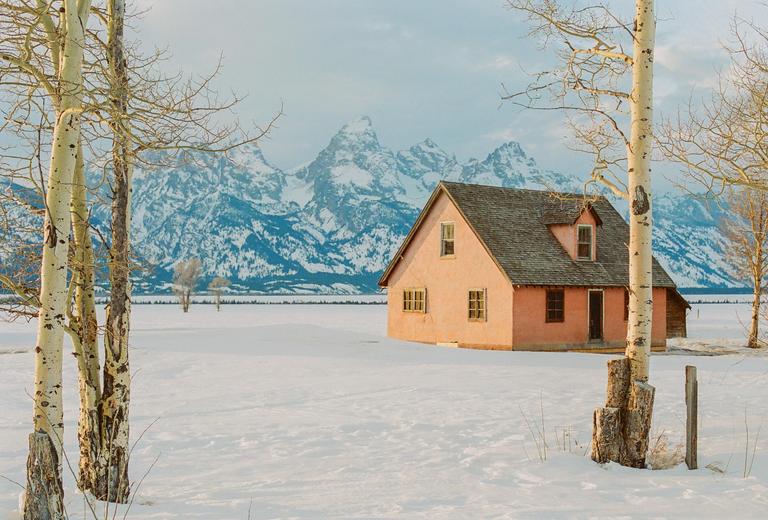 A pink house stands in a snowy field with mountains in the background and bare trees in the foreground.