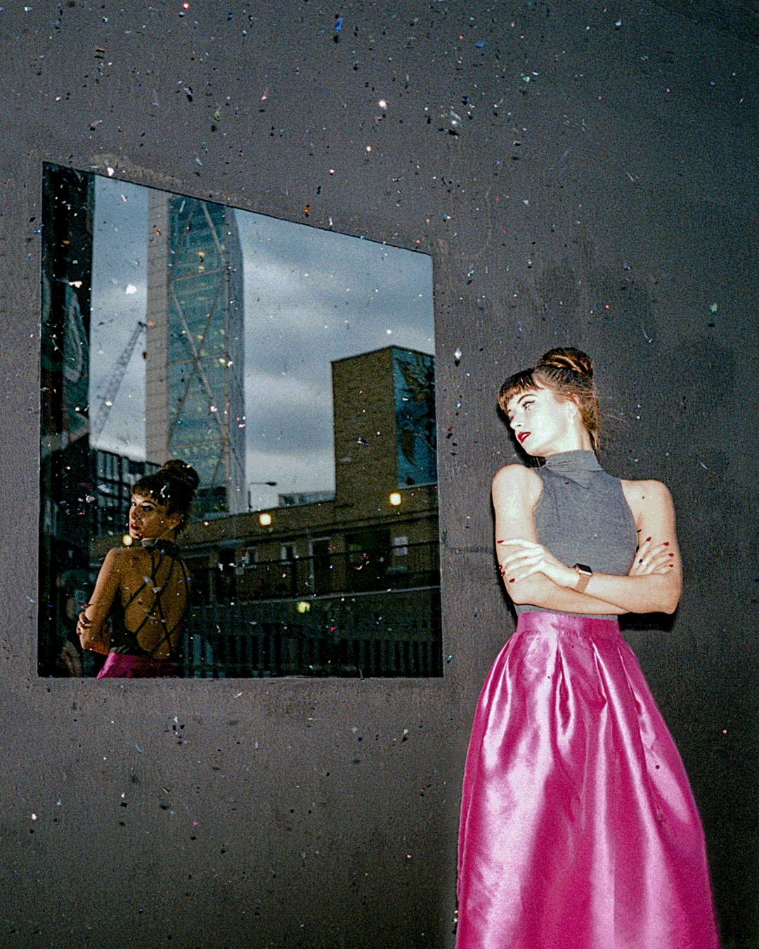 A woman in a pink skirt and grey top stands next to a mirror reflecting a city building, with glitter falling around her.