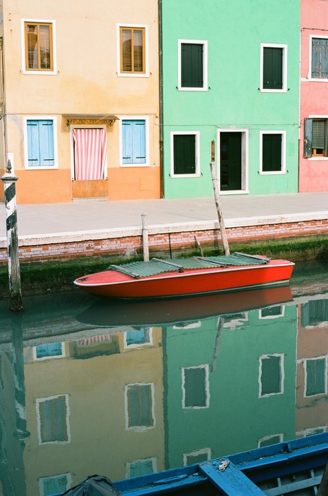 A red boat floats in a canal reflecting colorful buildings with windows and shutters.