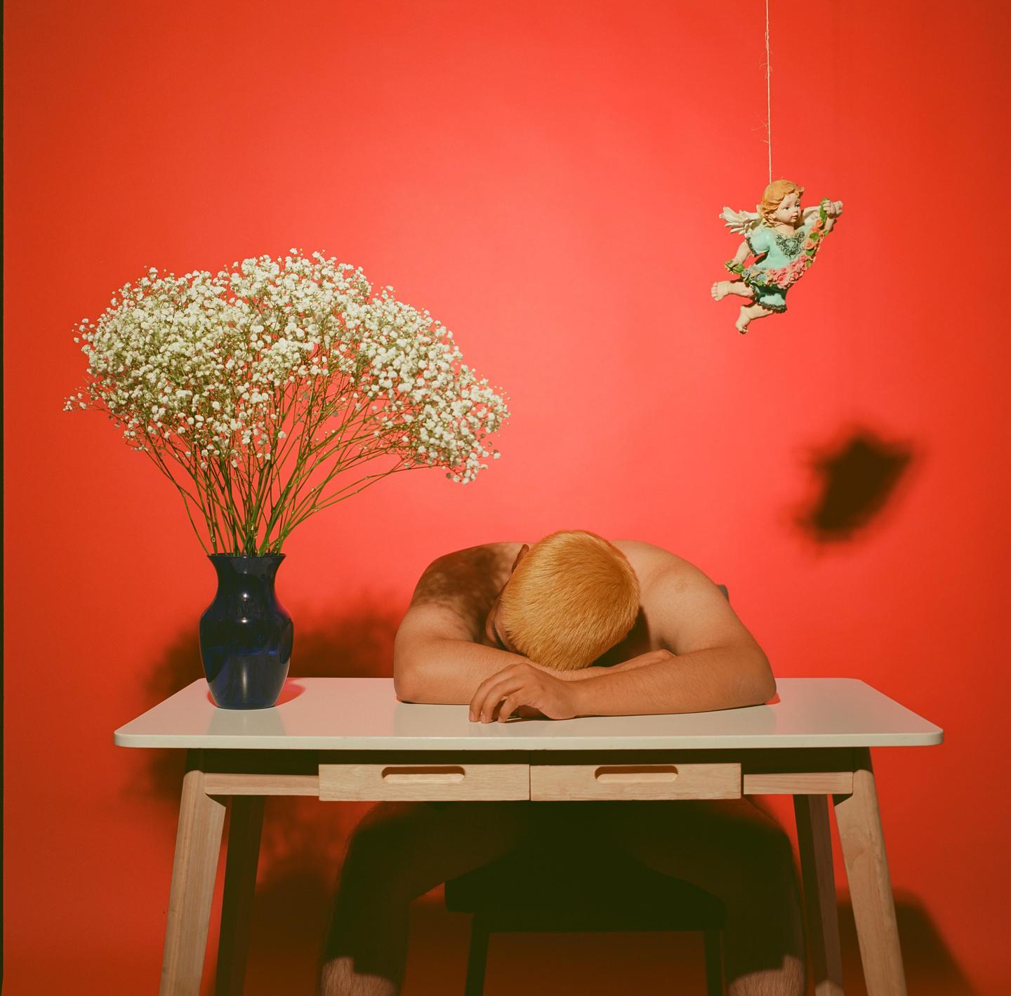 A man with blond hair rests his head on a table next to a vase of baby's breath flowers, with a cherub decoration hanging above him against a red wall.