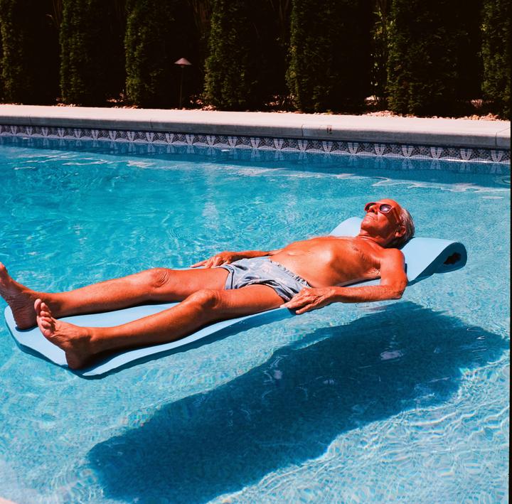 A man wearing sunglasses floats on a blue mat in a swimming pool.