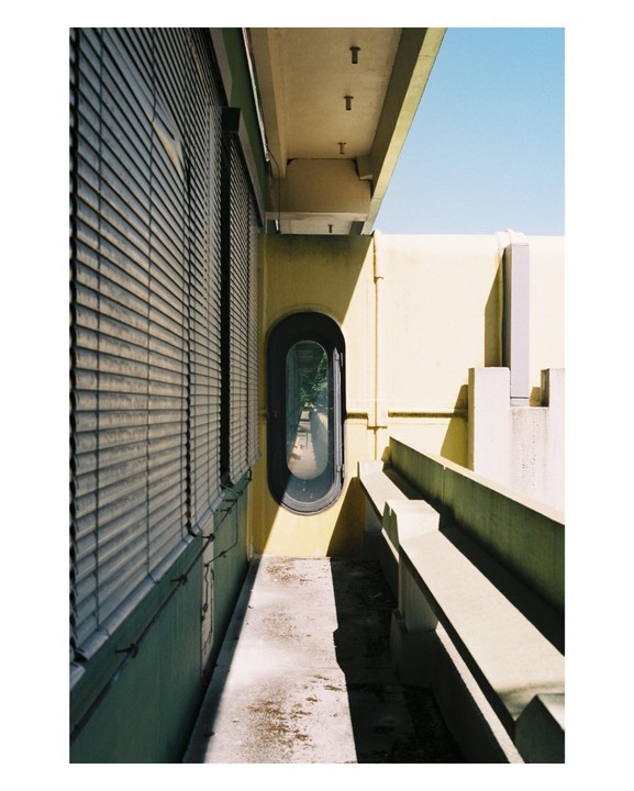 A narrow walkway with a long oval window on the right and a wall with horizontal blinds on the left.