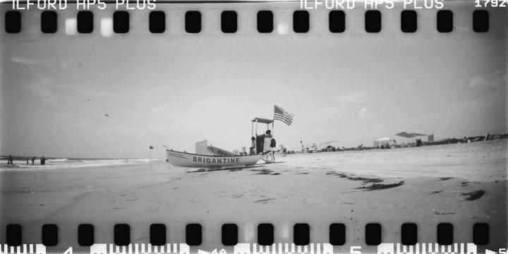 A lifeguard stand with an American flag and a boat named Brigantine are on a sandy beach with people and ocean waves in the distance.