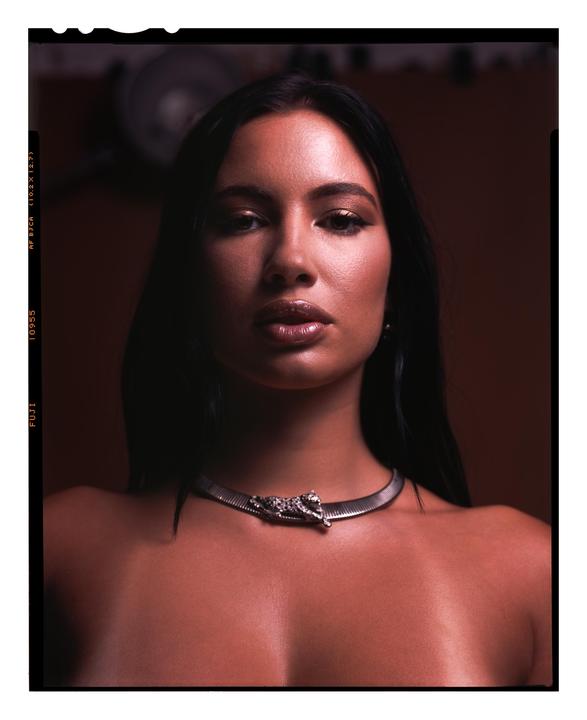 A woman with long dark hair and a metallic choker with a jaguar ornament looks directly at the viewer.