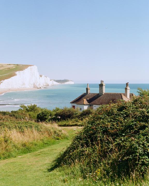 A white house with a brown roof is nestled among green bushes and grass, overlooking a beach with white cliffs and blue water.